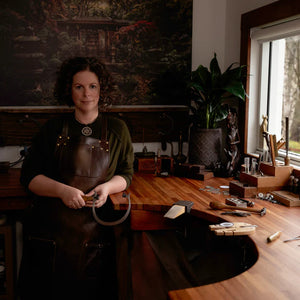 Jeweler Jean Burgers standing next to her jewelry bench in Emerald, VIC Australia. Holding a jewelers saw in her hands, wearing a leather apron. 